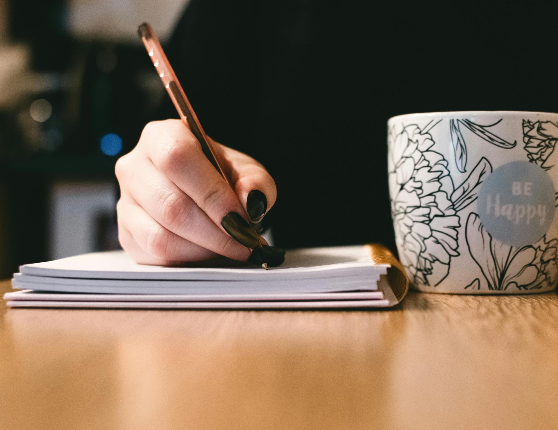Person writing in a notebook with a floral ceramic mug on a wooden desk.
