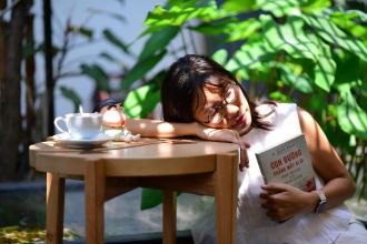 A woman sleeps on a table outdoors, holding a book, with a cup and toy nearby.