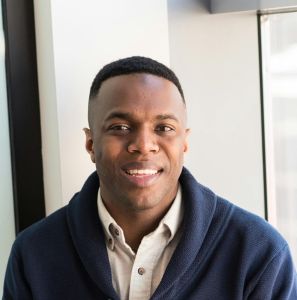 A professional black man smiling while seated by a window, embodying modern business vibes.
