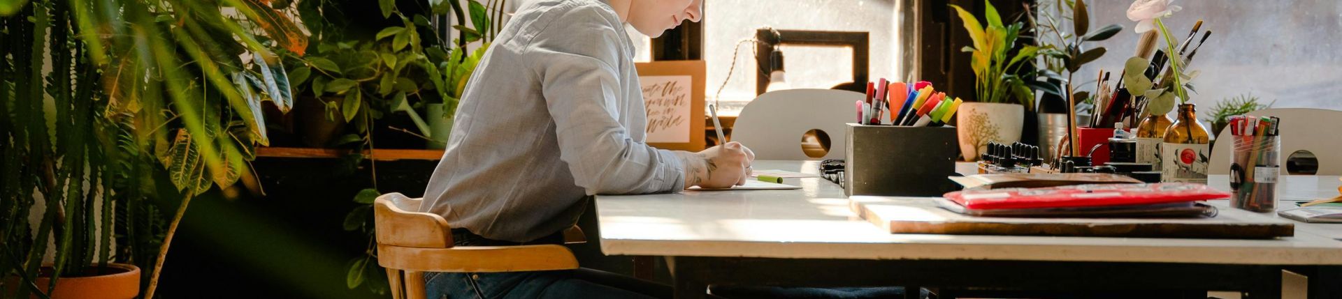 A female artist working diligently in a sunlit studio surrounded by plants and art supplies.