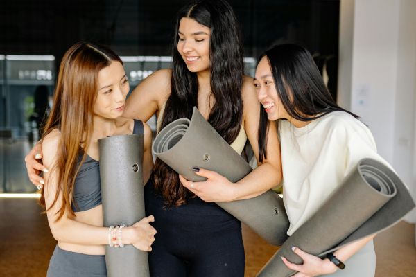Three women smiling and holding yoga mats during a fitness class indoors, embracing friendship and wellness.