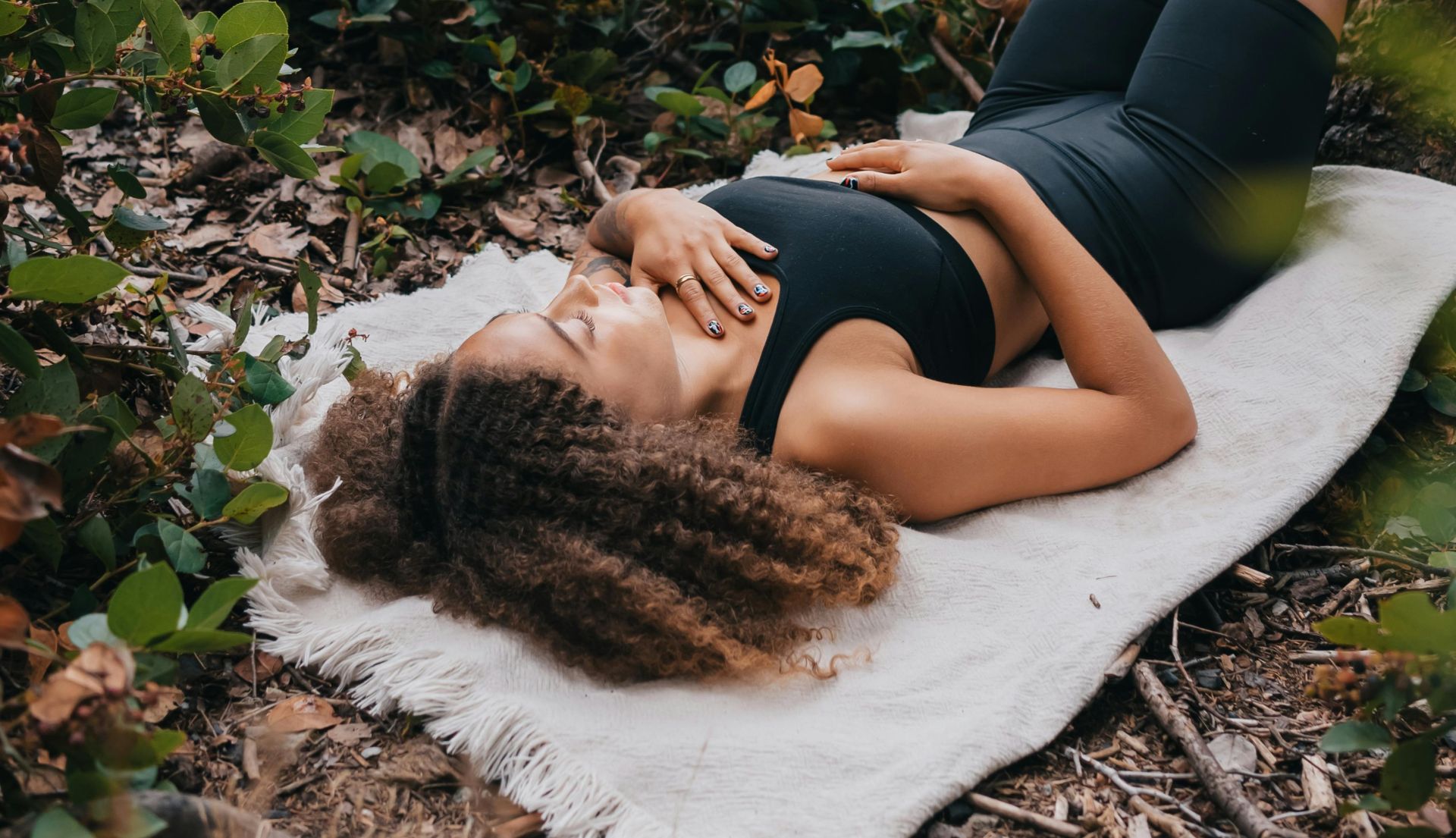 A woman with curly hair lying on a blanket outdoors in activewear, embodying relaxation and connection to nature.