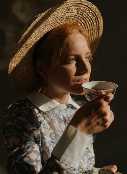 A redheaded woman in a straw hat enjoys chamomile tea in a porcelain cup.