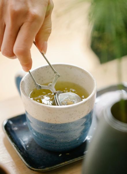 A hand using an infuser to steep green tea in a ceramic cup on a tray.